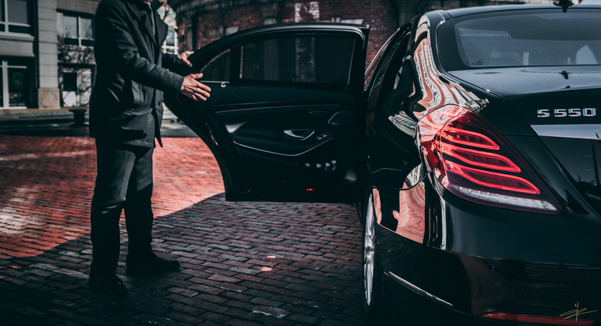 A professional chauffeur in a black coat opening the door of a luxury black Mercedes-Benz S-Class sedan, highlighting corporate events transportation and Clayton car service.