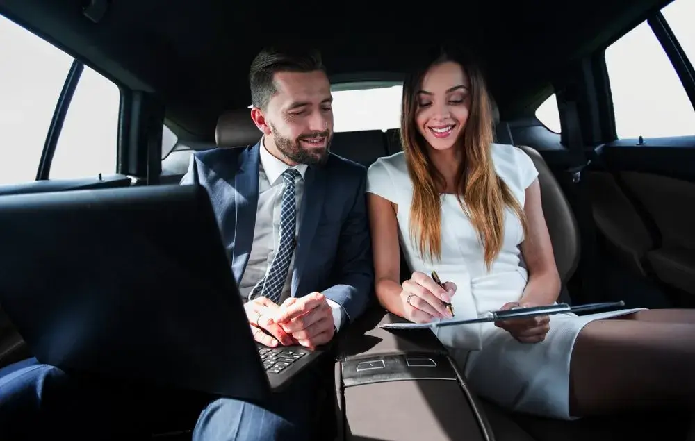 City to City Transfers City-To-City Transfers A man and a woman dressed in business attire sit in the back seat of a limousine, smiling while reviewing documents together on a clipboard and laptop