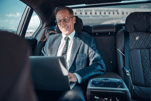 Our Blogs 10 A professional businessman in a suit and glasses smiling while working on a laptop in the backseat of a luxury black car, representing Gateway Limousine's executive transportation and St. Louis airport car services.